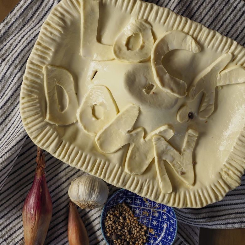 Photo of an uncooked puff-pastry pie with the words Lockdown as decoration - Pie is in a dish places on a tee towel with garlic, shallots and mustard seeds in a small dish alongside.