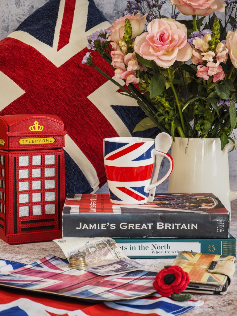 Photograph of items that represent Great Britain and the Union Jack.  Jamie Oliver's Great Britain Cook Book. Red Telephone Box Bag, Union Jack Mug and Teaspoon and Pink Roses in a Jug. REd Poppy and Mouse Mat.  Still Life Photograph to celebrate V.E. Day May 2020